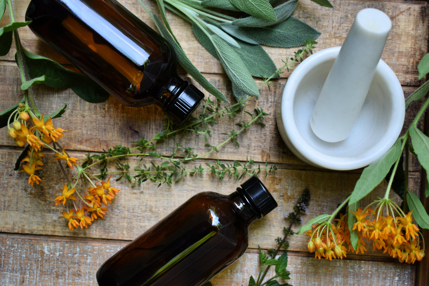 Two bottles of herbal extracts on a wooden surface surrounded by various herbs and flowers, with a mortar and pestle in the center.